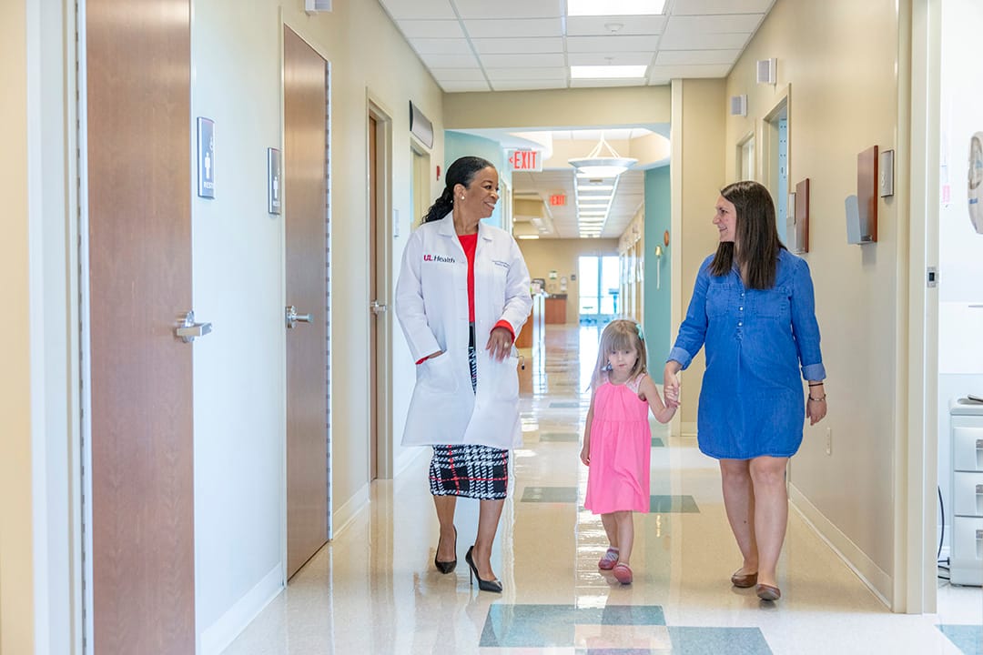 A doctor, walking with a mom and her daughter, explaining UofL Health's healthcare history in Louisville.
